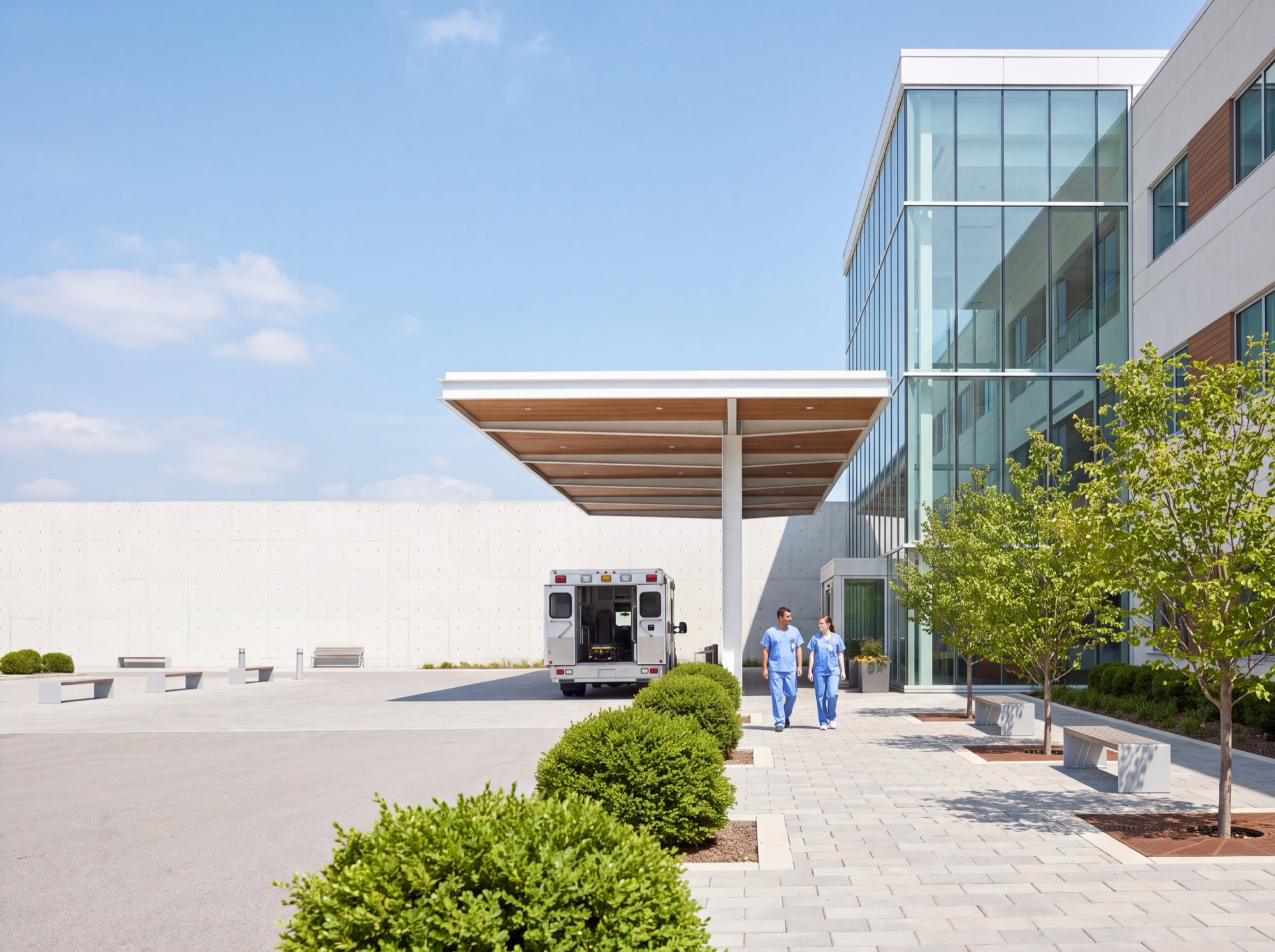 The entrance forecourt of a modern hospital: an ambulance waiting under the drop-off canopy and two staff in scrubs walking toward a glass atrium on a bright day.